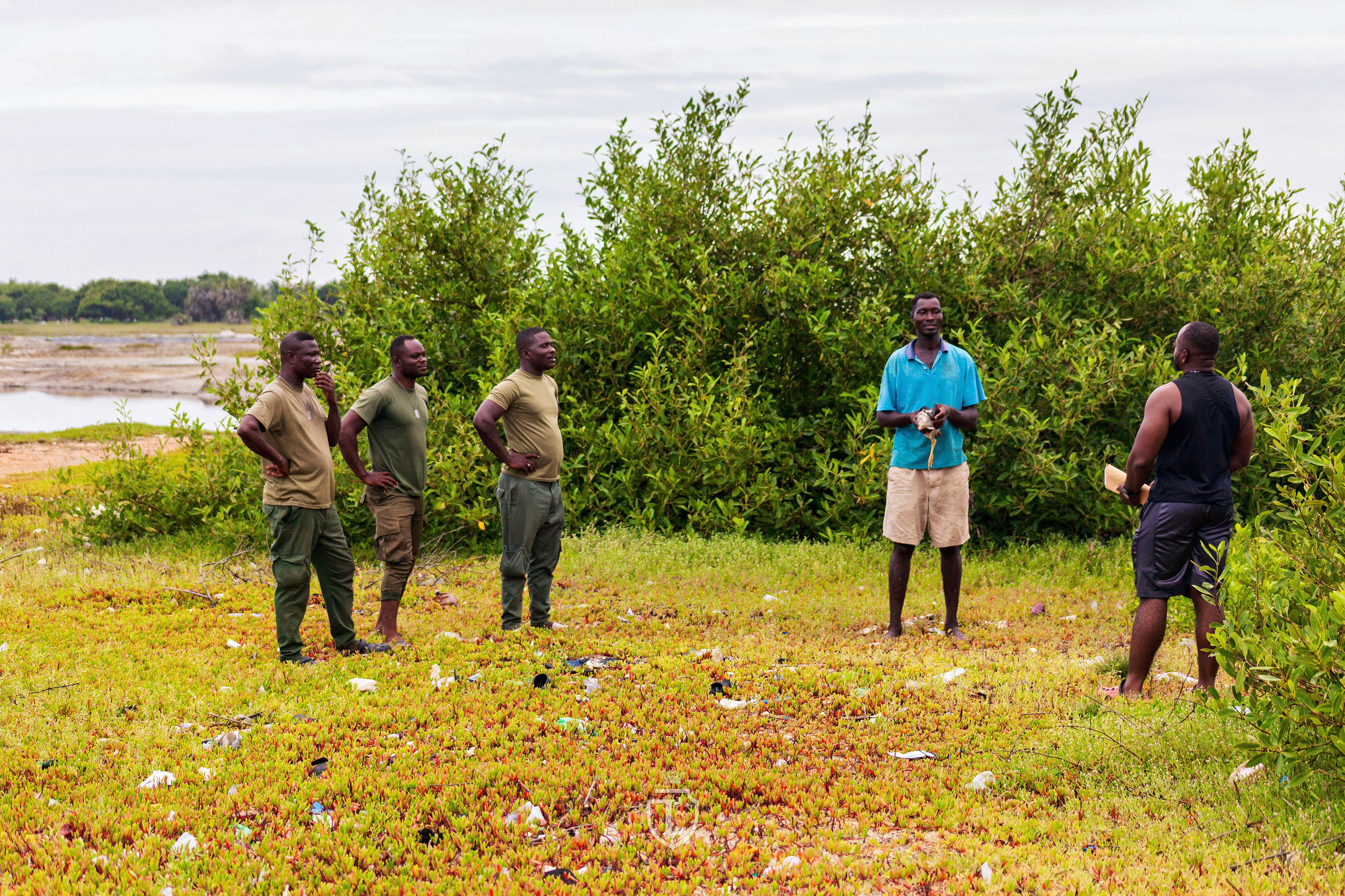 Wildlife officers and conservationists speak with a fisherman holding a heron, gesturing calmly while providing conservation education in a wetland landscape. © Alfred Vanderpuye of LV Studios