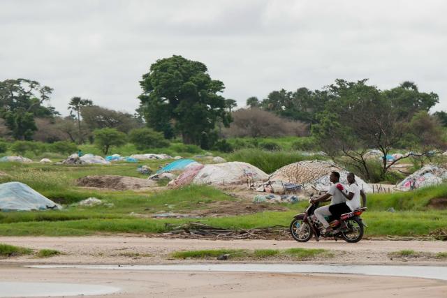 Senegal landscape photo showing two people riding a motorbike past green trees and some plastic waste in the background