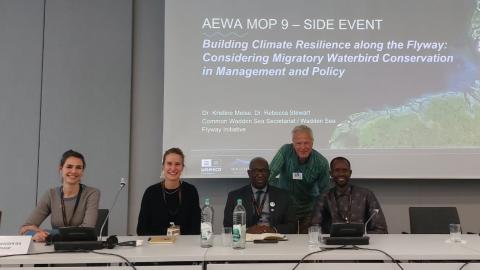 Five people sitting at a podium in front of presenter screen with the side event's title.