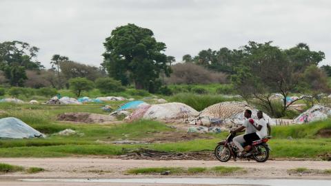 Senegal landscape photo showing two people riding a motorbike past green trees and some plastic waste in the background